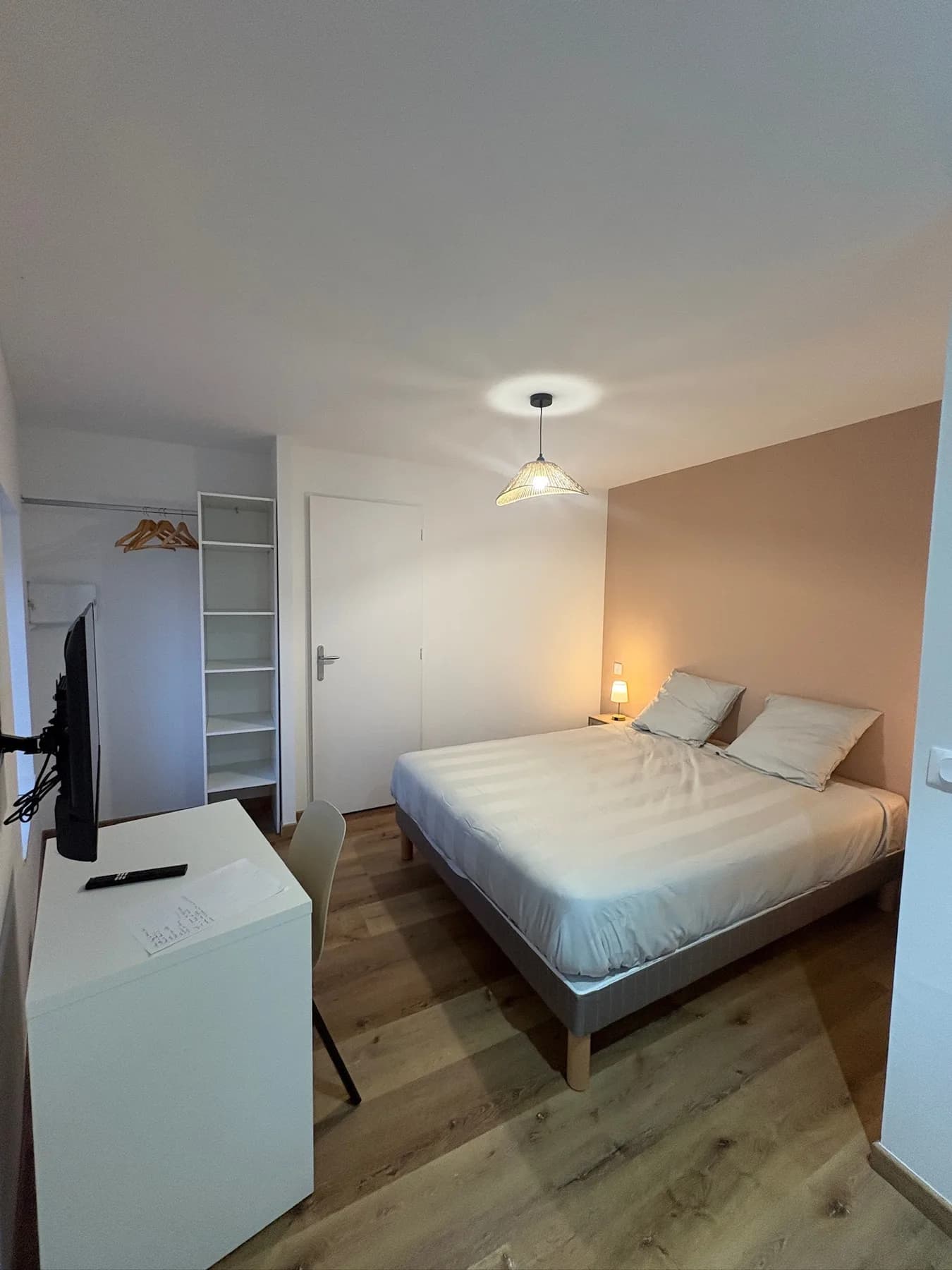 Chambre double avec linge de lit clair, bureau, télévision murale et penderie ouverte.